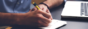 Hands writing notes near a laptop