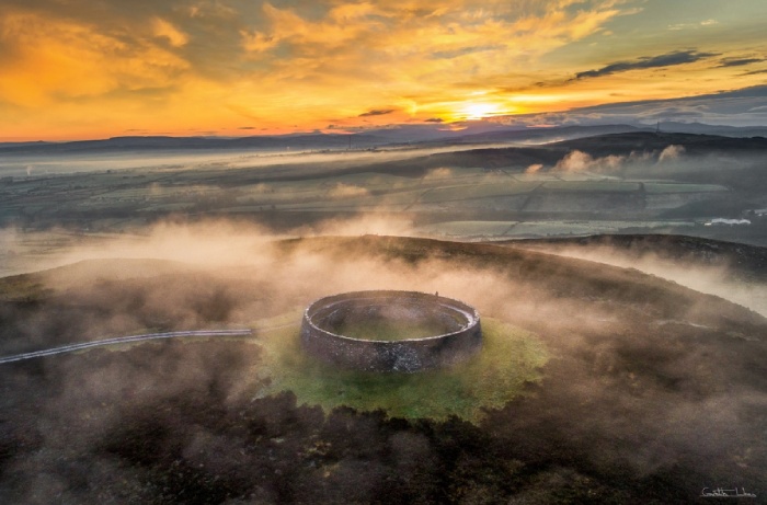 Aerial view of a circular stone fort on a hilltop surrounded by mist and illuminated by a colourful sunset.